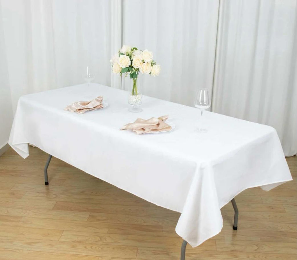 White tablecloth on a rectangular table with a vase of flowers and napkins against a white curtain background.