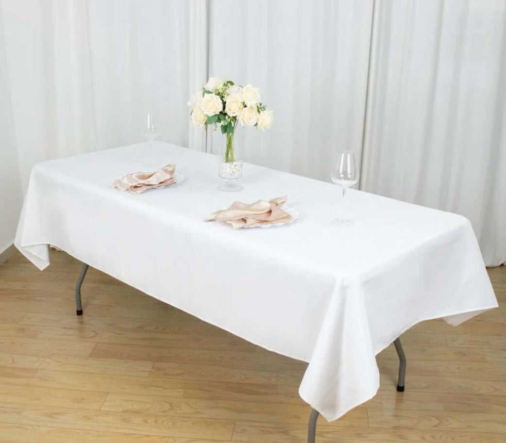 White tablecloth on a rectangular table with a vase of flowers and napkins against a white curtain background.